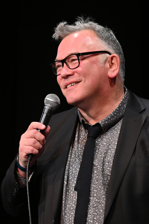 Stewart Lee holds a microphone, dressed in a grey shirt and black jacket, holding a microphone.