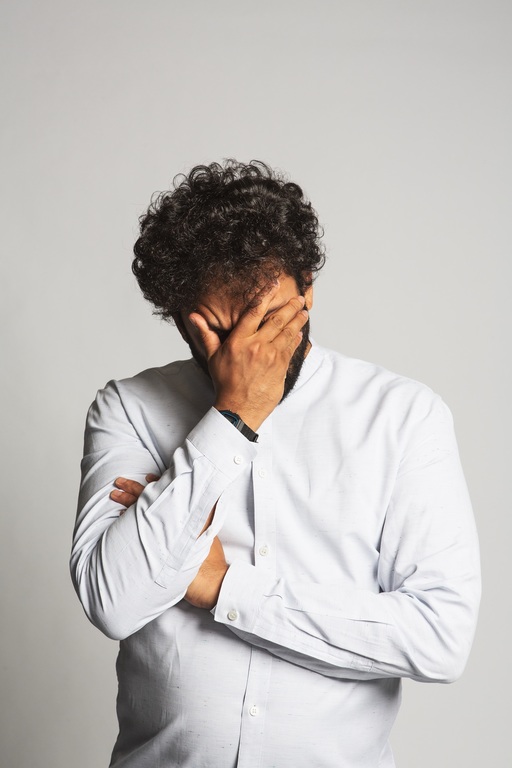 Nish Kumar, wearing a white shirt and holding his hand over his face.
