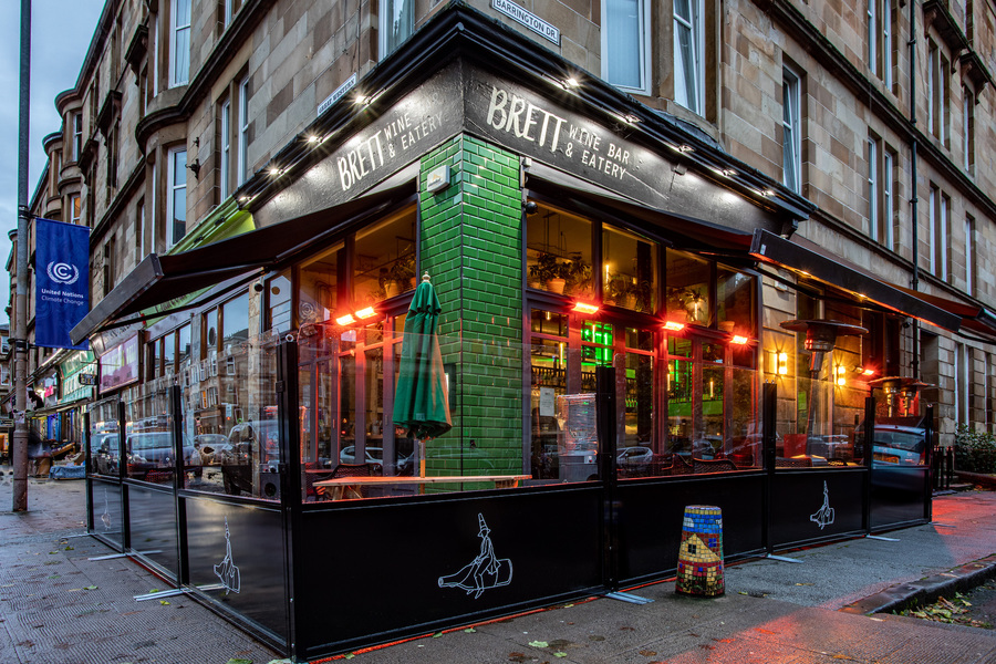 Exterior of photo of Brett bar in Glasgow. A corner bar with black awnings covering an outside seating area, and a green tiles corner nearest the camera. The tenement flats above are visible at the edges of the image.