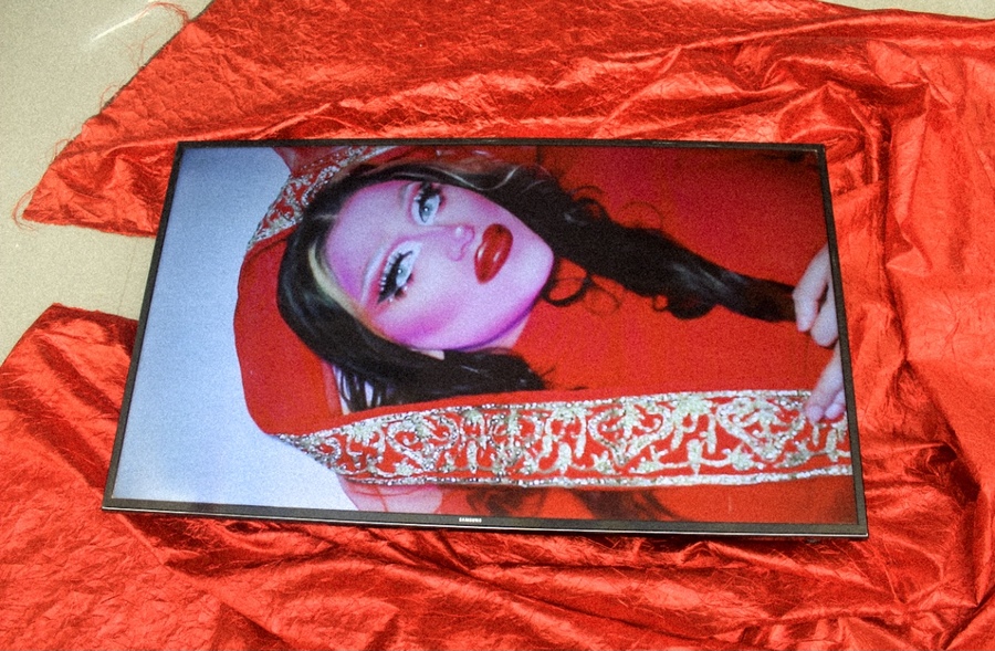 A television displays an image of a figure in bright, extravagant make-up wearing an ornate red outfit. The television sits on top of a piece of red fabric.