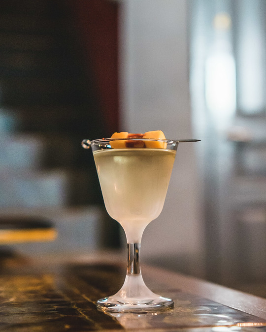 A clear cocktail with a yellow and red garnish draped across the mouth of the glass.