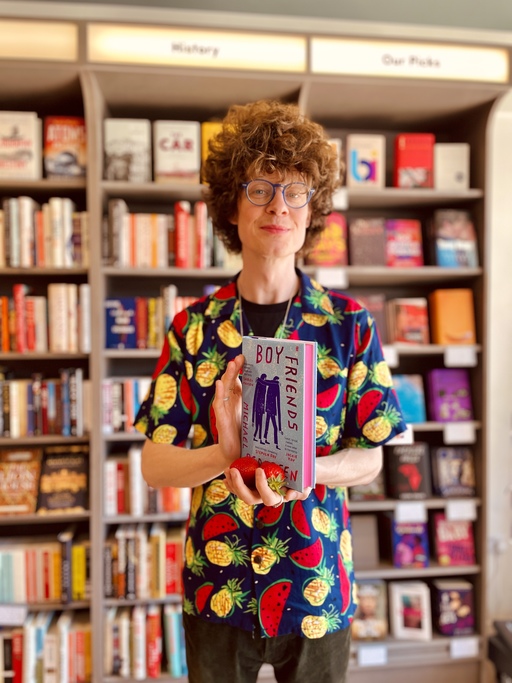 Michael Pedersen holds a copy of Boy Friends in front of a large bookcase. He is wearing a shirt with a repeating print of watermelons and pineapples.