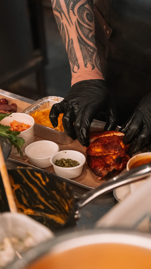 A tattooed arm wearing black food prep gloves places barbecued meat on a tray alongside a selection of small dishes.