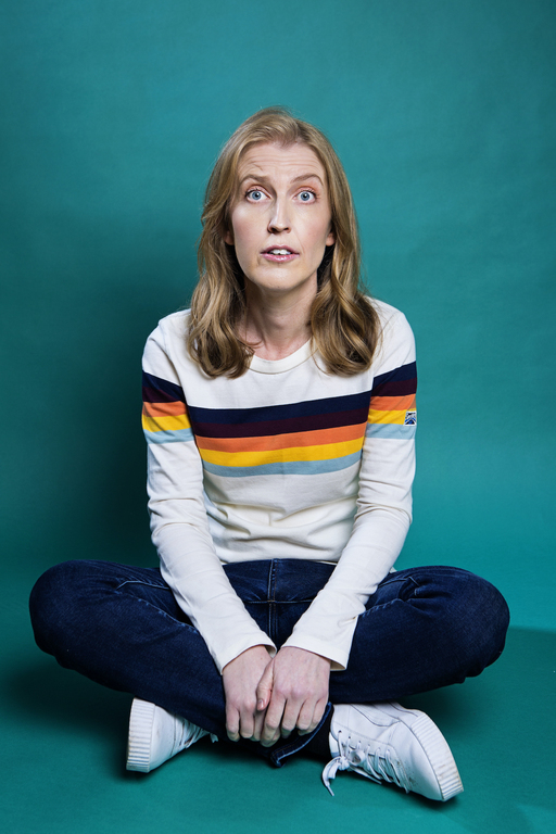 Comedian Heidi Regan sits crosslegged in front of a teal backdrop. She wears a white jumper with black, yellow, pink and blue stripes.