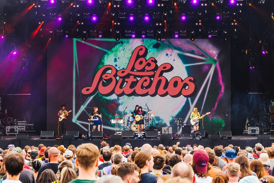 Los Bitchos performing on stage at Bluedot festival. The crowd can be seen in front of the band, with a large multicoloured banner reading 'Los Bitchos' visible behind.