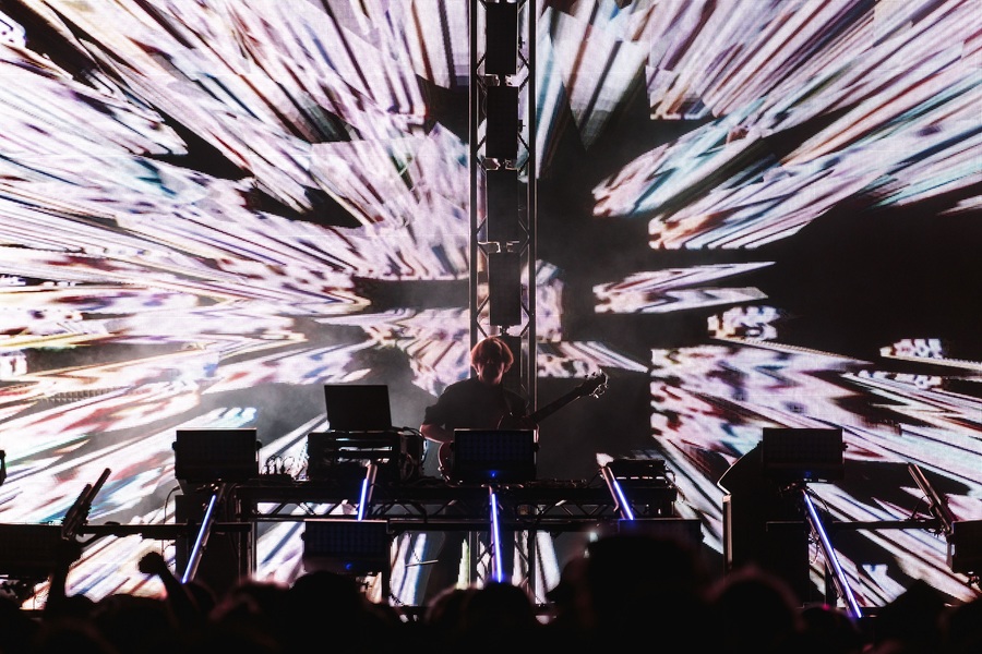 Squarepusher on stage at Bluedot. He stands behind a variety of laptops and synthesizers, with a large abstract visualisation behind.