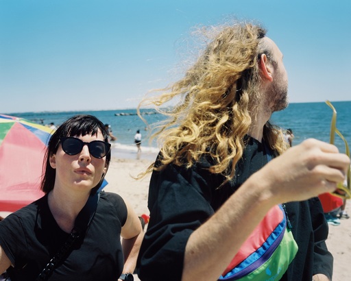 Sylvan Esso stand on a beach in front of umbrellas and people walking on the sand. Amelia Meath looks at the camera; Nick Sanborn is turned away.