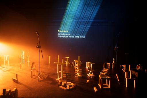 A microphone stand sits on a stage covered with wooden blocks arranged in a variety of configurations; some resemble office furniture. Text projected on the wall reads 'The audience: As the lights rise the city hums with the sound of work'