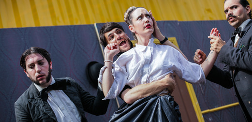 A scene from The Importance of Being... Earnest? A woman in a shirt and dress is held from behind by a man in a tuxedo, while two other men stand nearby.