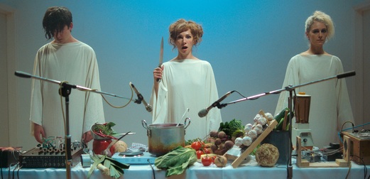 Still from Flux Gourmet. Three figures in long white robes stand behind a table filled with vegetables and cooking equipment, with two microphones pointed at a saucepan. The central figure holds a knife.