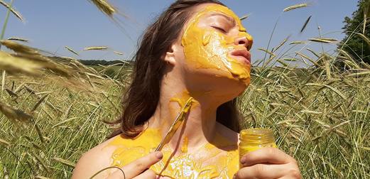 A woman, smearing mustard on her face and shoulders with a butter knife, while standing in a field of wheat.
