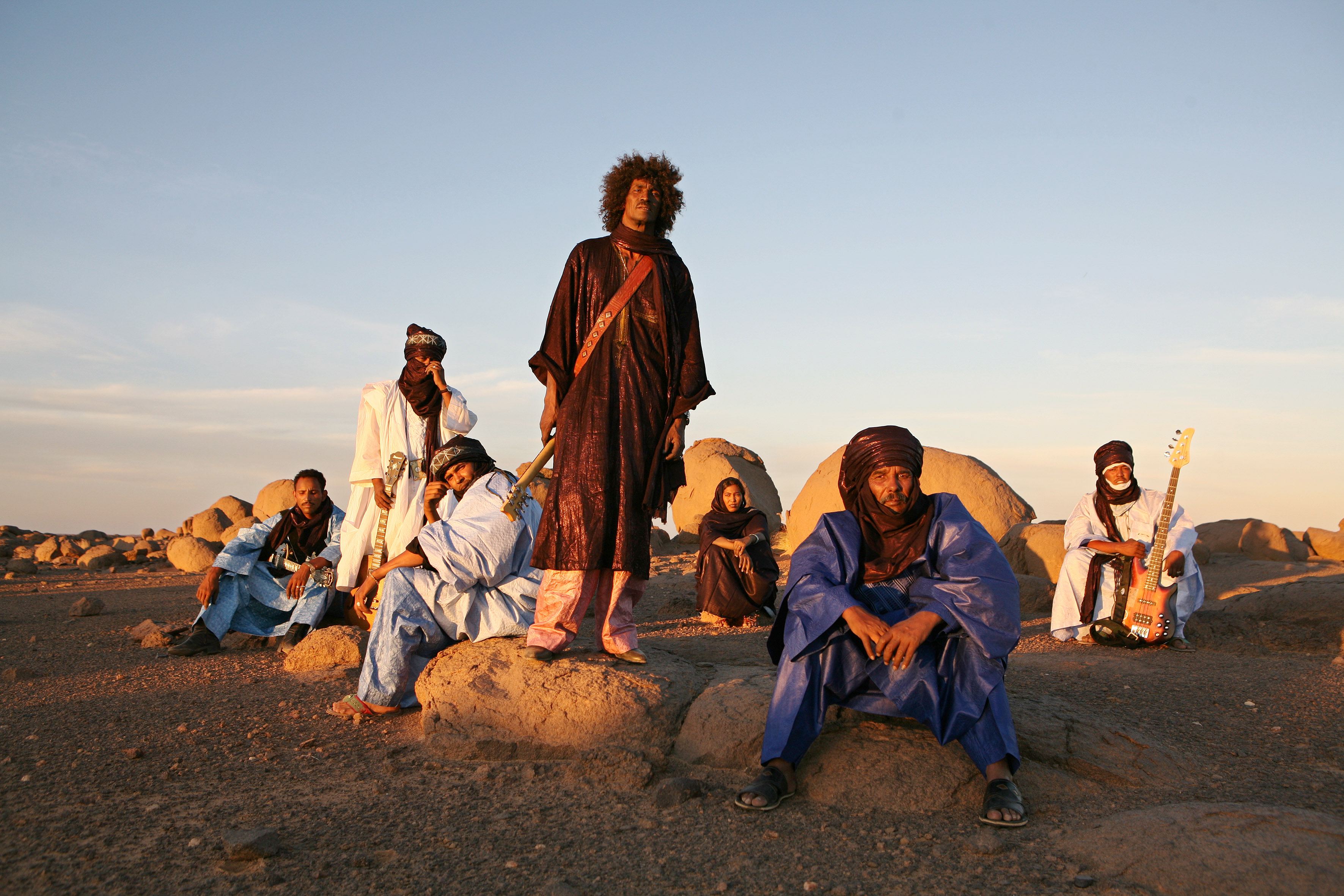 The members of Tinariwen, some sitting and some standing, each wearing long flowing robes. They pose against a desert scene populated by large rocks