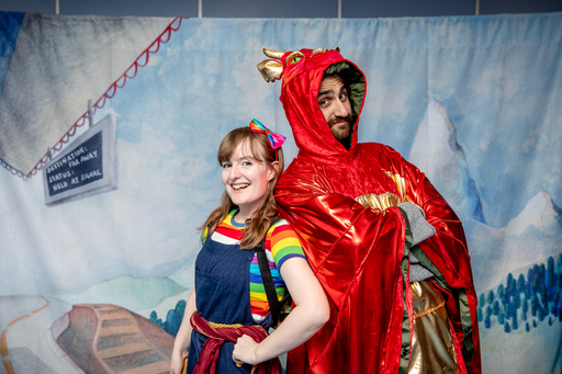 Two people – on wearing a rainbow-striped top and dungarees, another dressed in a red dragon costume, stand back to back in front of a painted backdrop.