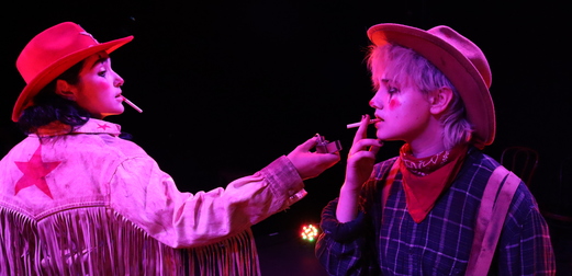 Two rodeo clowns stand under a purple light, one offering the other a light for their cigarette.