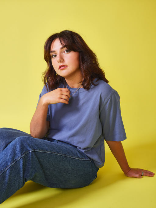 Ania Magliano, wearing a blue top and blue jeans, sitting in front of a yellow background.
