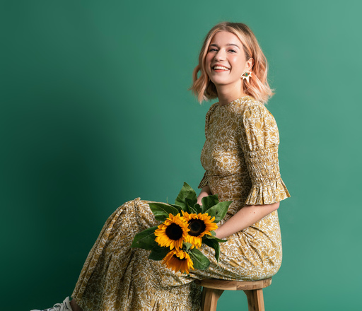 Olga Koch in a beige patterned dress, holding a bunch of sunflowers, in front of a green background.