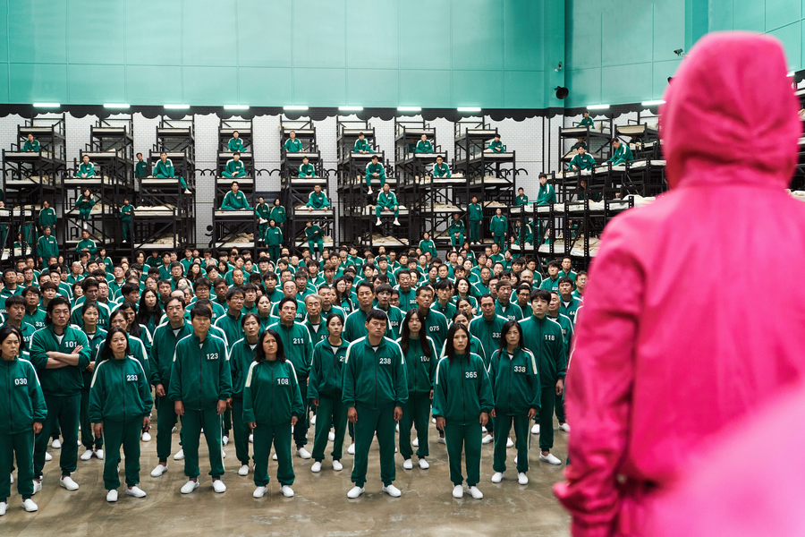 A still from Squid Game. A hooded figure dressed all in pink stands in front of a large crowd of people dressed in identical teal tracksuits.
