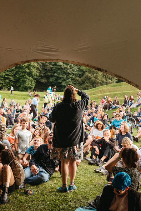 Josie Long performing at Jupiter Artland. She stands in the doorway of a tent, in front of a crowd assembled on the grass.