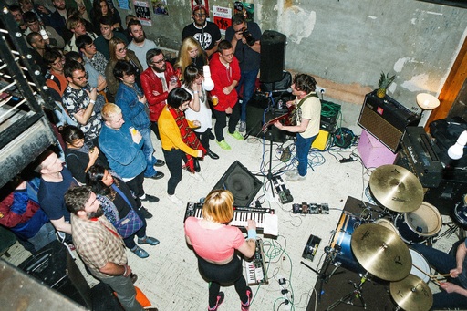 A band performs in front of a small crowd. A keyboardist, guitarist and drummer are visible, with a selection of pedals on the floor around the band.