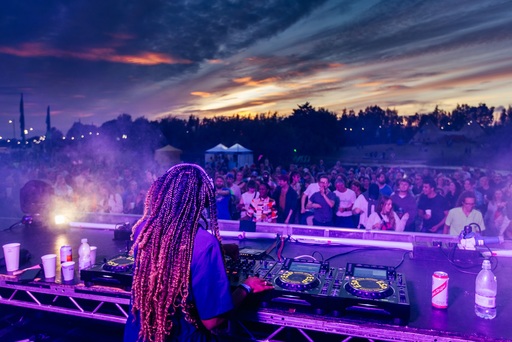 Jamz Supernova standing behind a set of turntables, with a crowd in front of her and a sunset behind them.