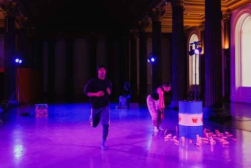 A scene from Ashanti Harris' An Exercise in Exorcism. A man runs across a hall, while a woman arranges a selection of blocky objects in the background.