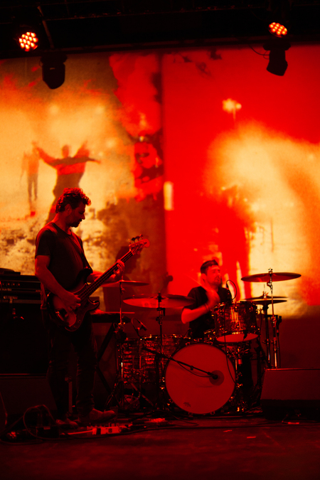 Two members of Godspeed You! Black Emperor on stage in front of a red and orange abstract projection. One plays the guitar, while the other plays the drums.