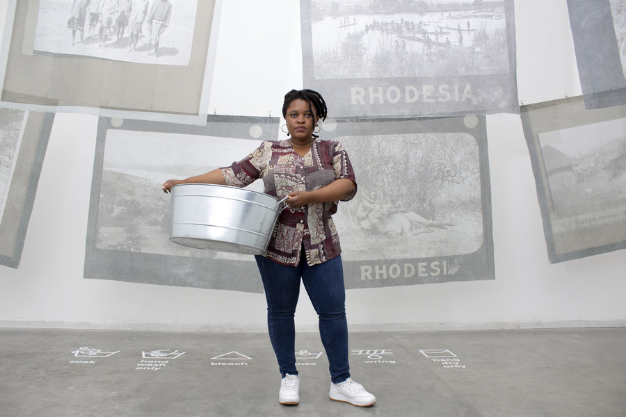 Tanatsei Gambura stands in front of a collection of large-scale black and white images. She is holding a washing pail.