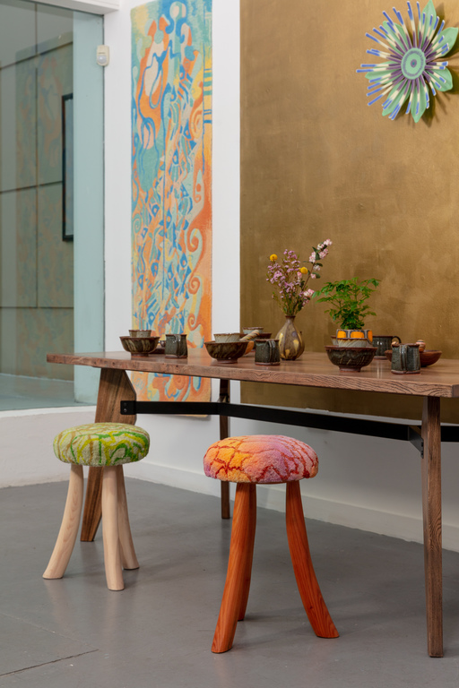 Exhibition view of Crafthouse. Two stools with tufted cushions, a table with a variety of bowls and cups, and two wall hangings.