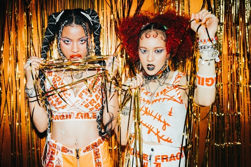 Nova Twins stand in front of a gold curtain; they are wearing 'Fragile' parcel tape across their bodies.