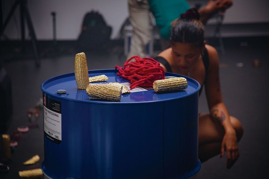 A collection of gold-plated corn cobs sit on top of a blue oil drum. A dancer crouches behind.