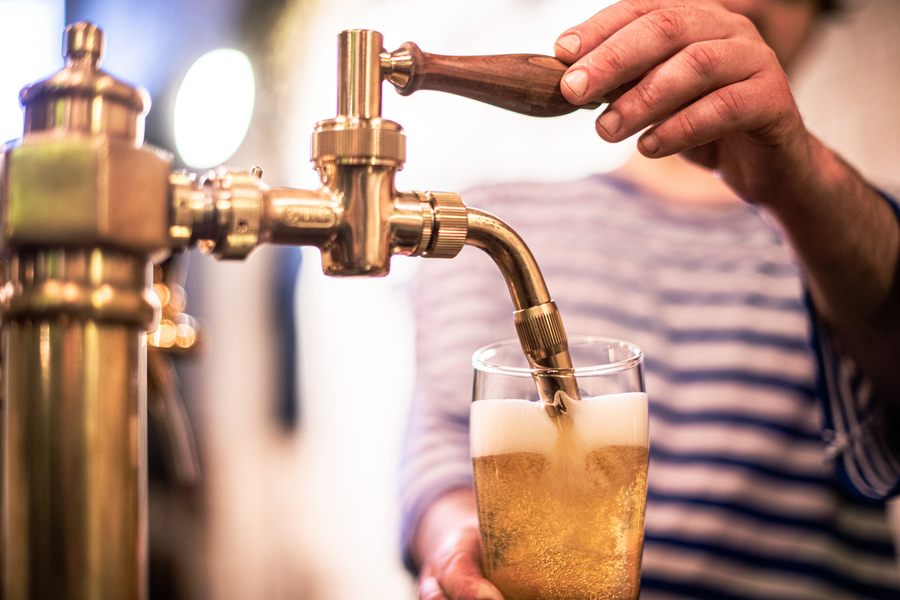 A pint of beer being poured from a golden tap. The barman wears a blue and white-striped T-shirt