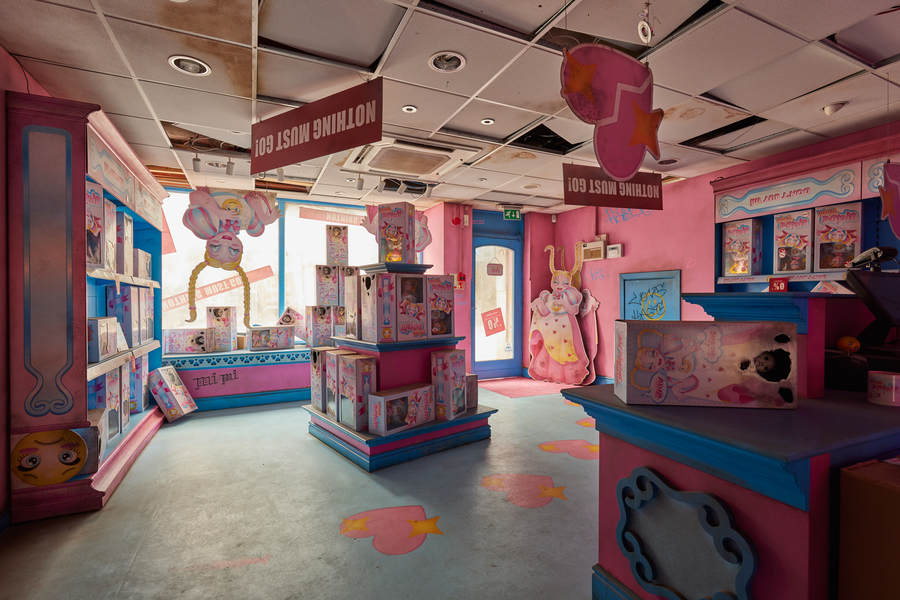 Installation view of Rachel Maclean's Mimi. A mocked-up shopfront with upside-down signage, items strewn on the floor and large artworks depicting a doll with long blonde hair.