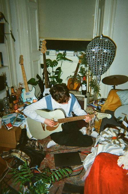 Carsick Charlie, playing guitar crosslegged on the floor. Various items are dotted around the room.