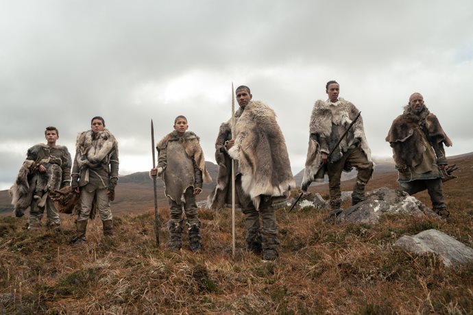Still from The Origin. A group of men wearing animal fur and holding spears stand on a hill