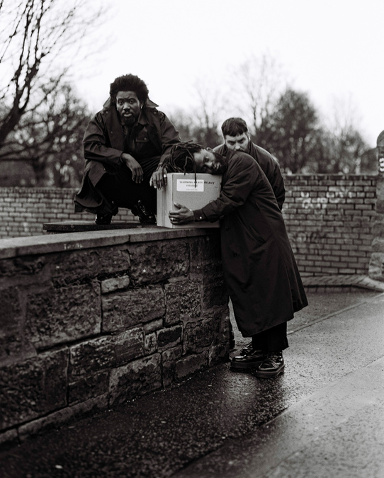 A black and white photo of Young Fathers. Kayus leans on a large box with the words 'Heavy Heavy' written on the side; Alloysious crouches on top of a short wall, with Graham standing behind.