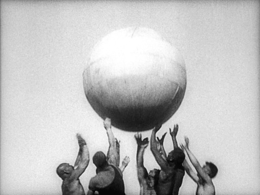 A still from In Spring; a black and white image group of men pushing a large ball into the air.