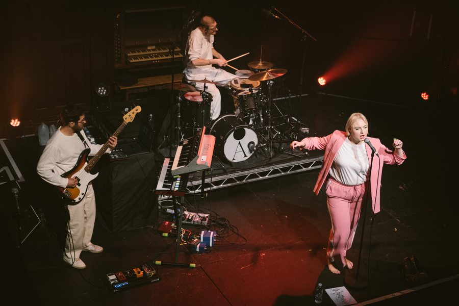 Lapsley on stage in Glasgow. She is singing into a microphone, wearing a pink suit and white shirt. A guitarist and drummer, dressed all in white, can also be seen on stage.