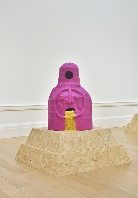 A bright pink beehive with carved features to resemble a human face. The face appears to be vomiting beeswax, and the hive sits on a plywood plinth.