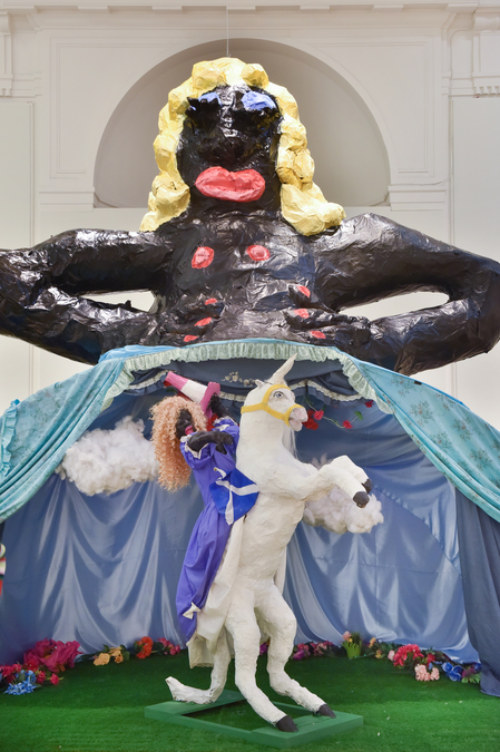 A large sculpture of a Black woman with blonde hair and an oversized skirt. In front of the large sculpture, A figure of a Black woman rides a unicorn while holding a traffic cone.