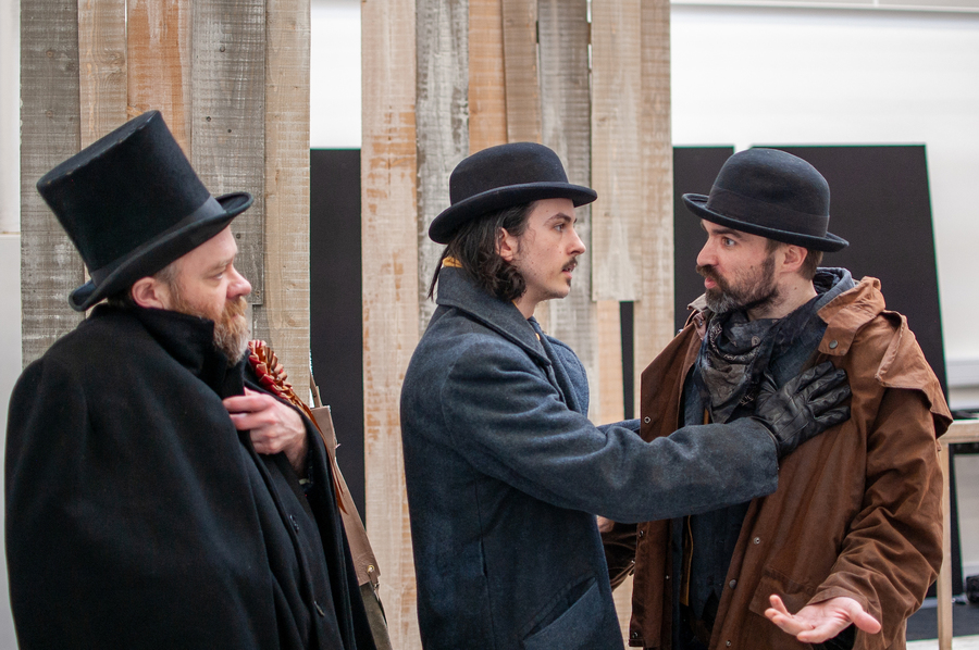 A photo from rehearsals for Stornoway, Quebec. Three men dressed in thick coats and black hats argue in front of a partially-built set.