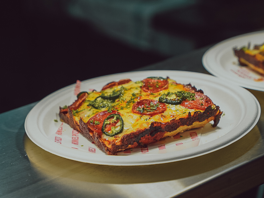 A square slice of pepperoni pizza on a white plate, sitting on a metal kitchen bench.