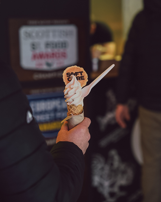 A soft serve ice cream with a 'Soft Core' branded wafer, held by a figure in a black jacket.