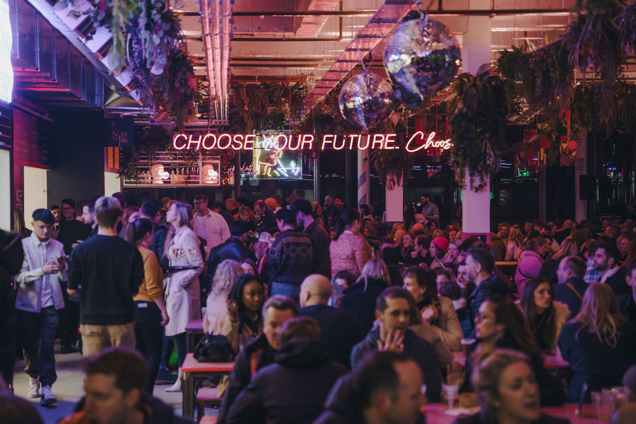 A large crowd sits at tables in Edinburgh Street Food. A pink neon sign, partially obscured by a plant, reads 'Choose Your Future, Choose'