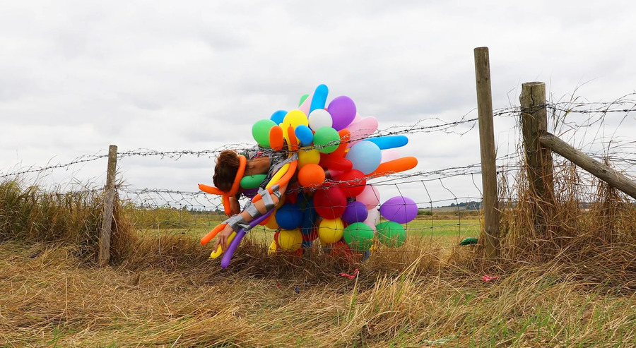 A figure covered in colourful balloons tries to squeeze through a wire fence in a field.