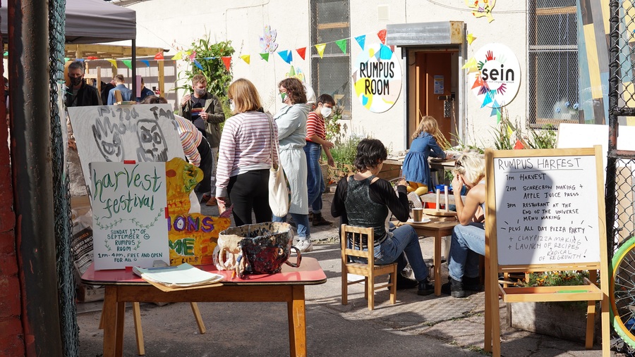 An exterior view of Rumpus Room. Two people sit at a small table, while others chat at the other side of the yard.