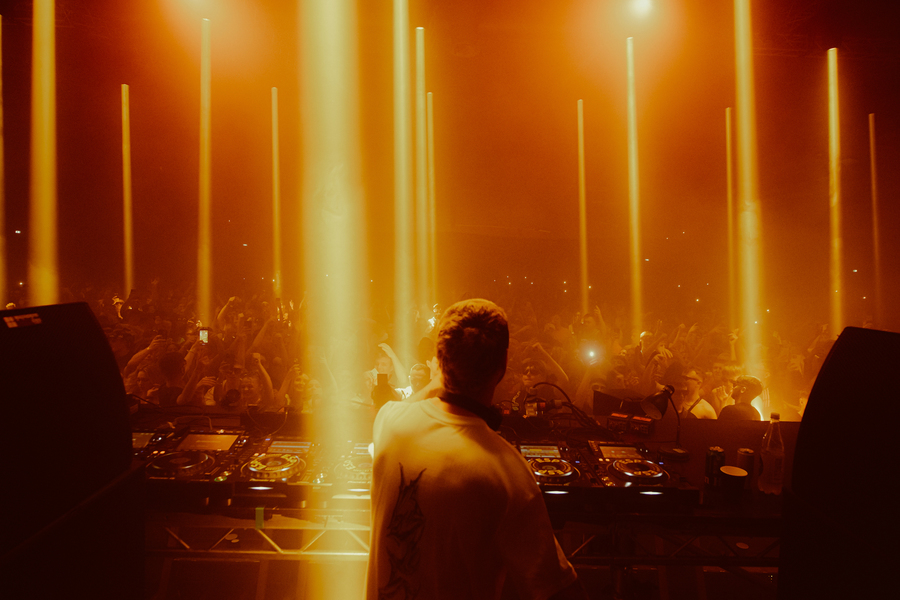 Franck on stage at Terminal V. The crowd can be seen beyond the DJ booth, with shafts of yellow light coming down from the ceiling.