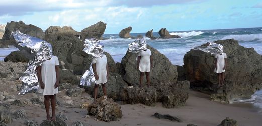 Four Black figures in white dresses, with tin foil headpieces, stand on rocks by a beach.