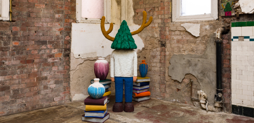 A figure with a tree-shaped head stands in the corner of a room, surrounded by books and vases.