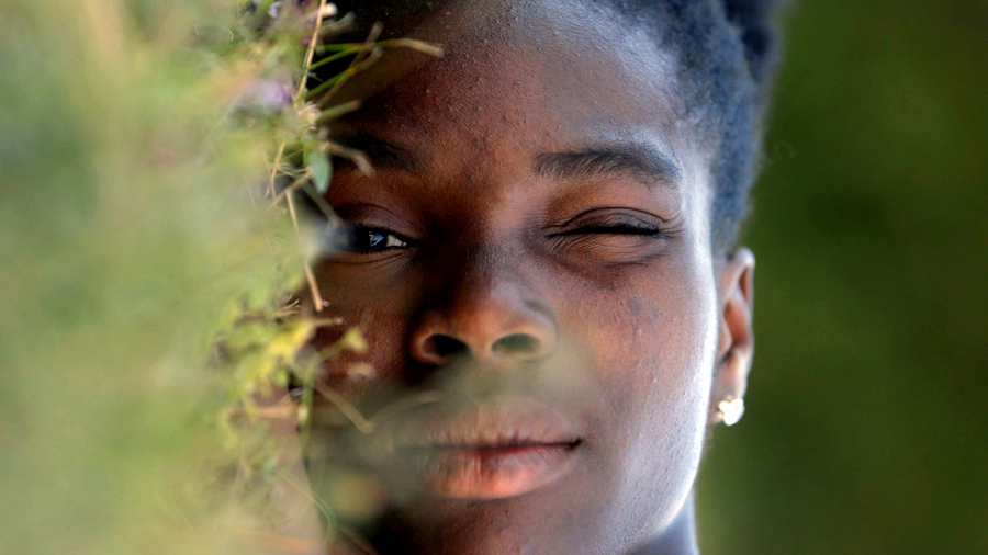 A still from the film This Place is a Message. A Black woman lies on the grass, with one eye closed.