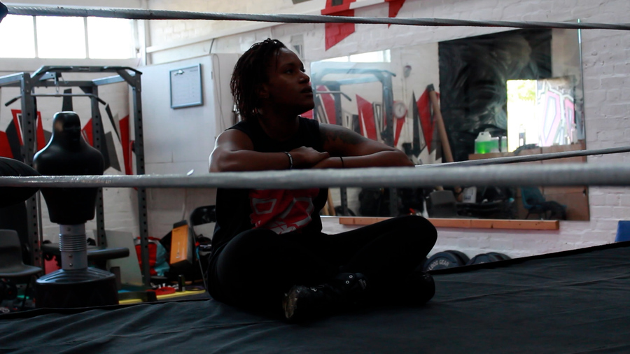 A Black wrestler sits cross-legged by the side of a wrestling ring, leaning her arms on one of the ring ropes.